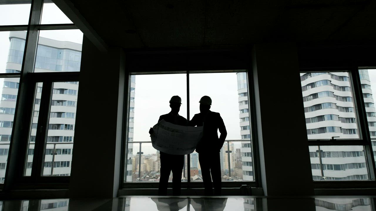 Two architects studying a blueprint in front of tall windows